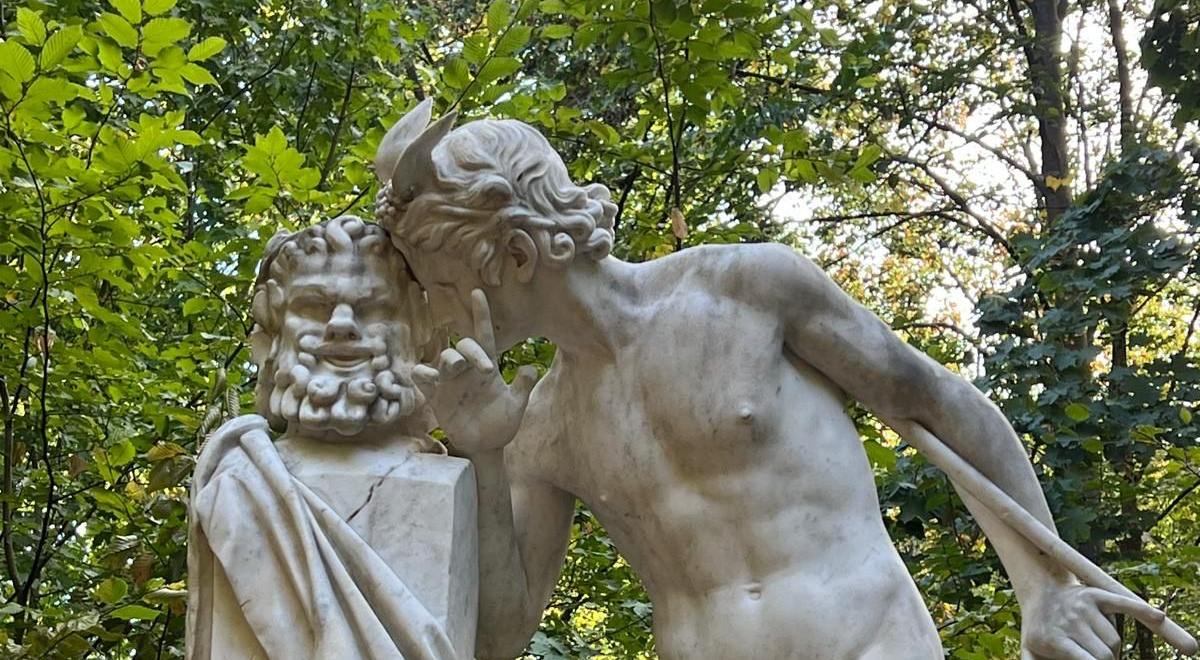 Statue de marbre : un jeune homme murmure à l'oreille d'un buste barbu, dans un sous-bois